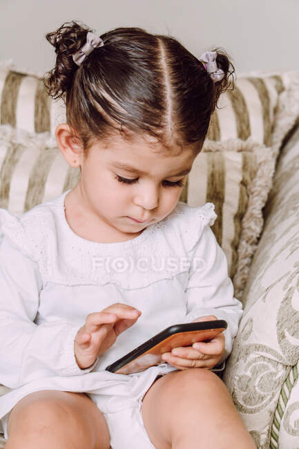 Criança adorável sentada no sofá em casa e assistindo desenhos animados interessantes no telefone móvel — Fotografia de Stock