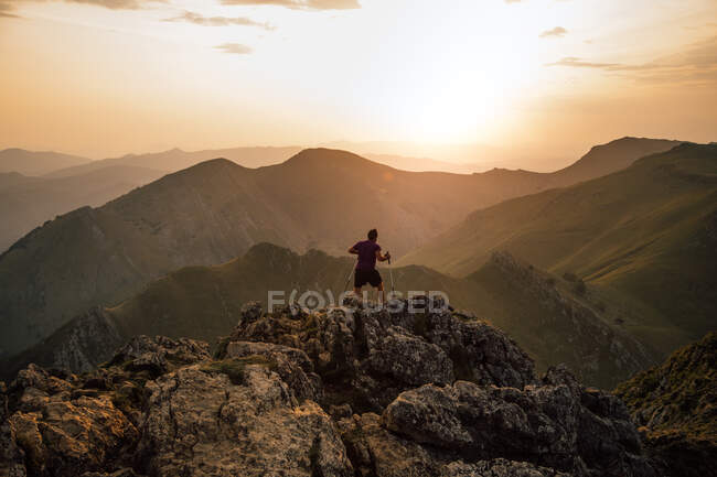 Vue arrière de l'homme bondissant sur les rochers du haut sommet de la crête de montagne sous un ciel nuageux au coucher du soleil — Photo de stock