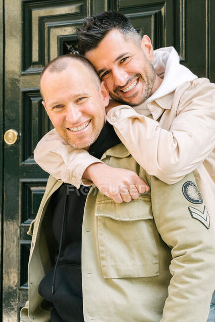 Cheerful young homosexual diverse men in stylish outfits smiling and embracing while standing on street near door and looking at camera - foto de stock