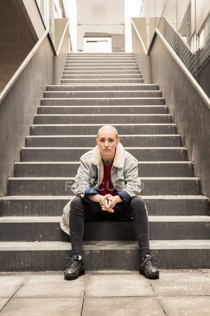 Junge transsexuelle Person in lässiger Kleidung sitzt auf einer Treppe zwischen Häuserwänden und blickt in die Kamera — Stockfoto