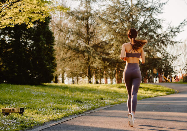Atleta donna con vista posteriore in abbigliamento sportivo che corre su strada asfaltata mentre distoglie lo sguardo durante l'allenamento cardio alla luce del sole — Foto stock