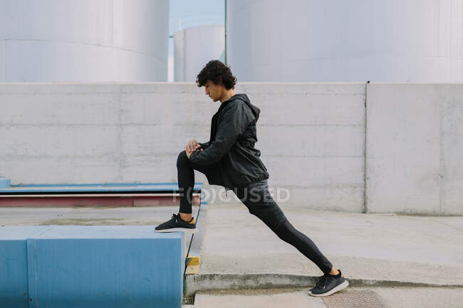 Vista lateral macho de corpo inteiro em roupas esportivas pretas totais inclinando-se com a perna na doca enquanto se alonga no distrito industrial da cidade — Fotografia de Stock