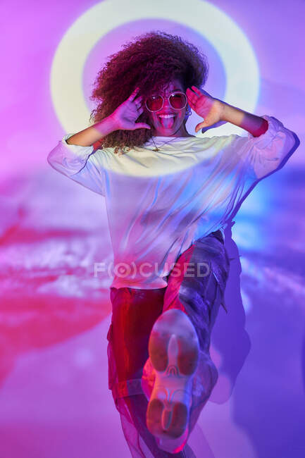 Fiduciosa giovane ballerina afro-americana felice in abbigliamento informale e occhiali da sole che balla puntando i piedi verso la fotocamera facendo volti in luci al neon in studio — Foto stock
