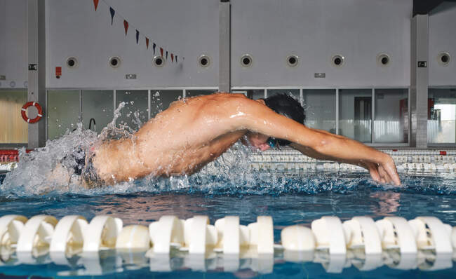Вид сбоку сильного мужчины, плавающего в купальной шапке и совершающего удар бабочкой во время тренировки в бассейне с голубой водой — стоковое фото