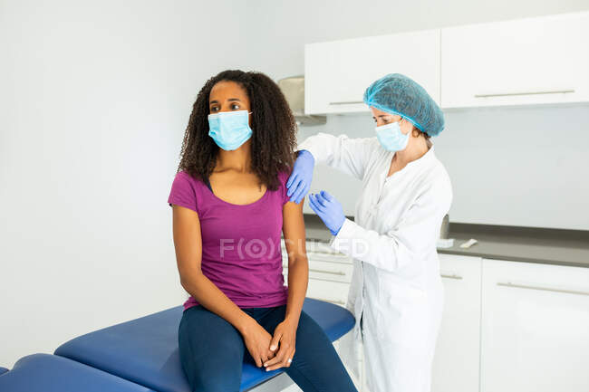 Spécialiste féminine en uniforme de protection, gants en latex et masque facial vaccinant la patiente afro-américaine en clinique pendant l'épidémie de coronavirus — Photo de stock