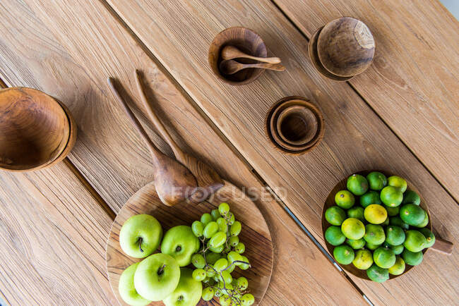 Top view of fresh ripe green apples with grapes placed on wooden tray near plate of limes and various traditional bowls served on table in sunlight — Stock Photo