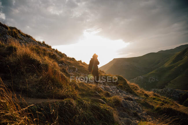 Side view of female walking downhill on rocky slope of mountain in green valley with cloudy sunset sky on background — Stock Photo