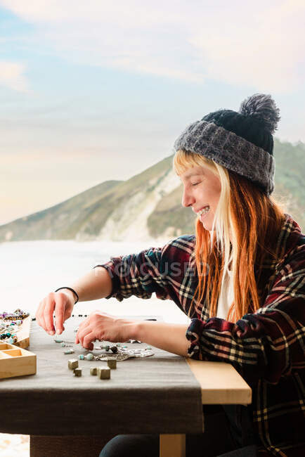 Vista lateral de feliz contenido viajero femenino creando accesorios hechos a mano mientras está sentado en la mesa de madera en el camión estacionado en la playa - foto de stock