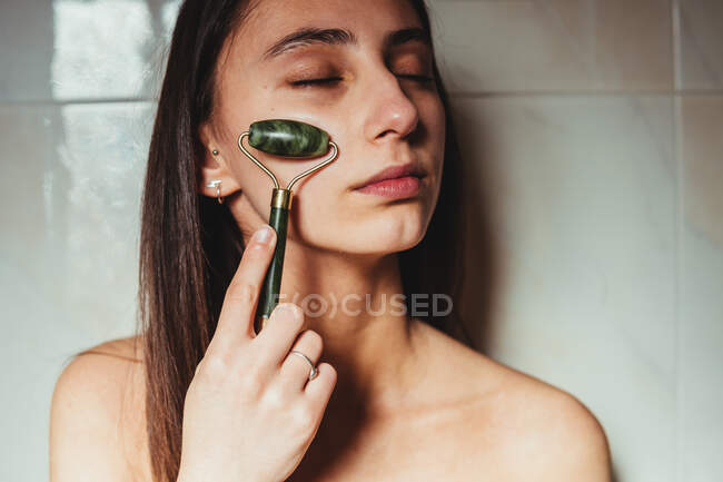 Концентрированная молодая женщина с закрытыми глазами массирует щеку нефритовым роликом о керамическую стену — стоковое фото