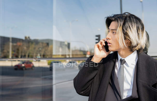 Молодой хорошо одетый этнический предприниматель мужчина с окрашенными волосами разговаривает по мобильному телефону, глядя в сторону стеклянной стены и городской дороги — стоковое фото
