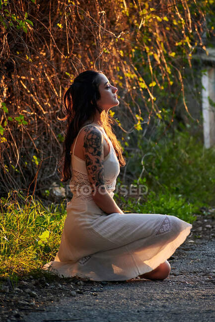 Dreaming lady with tattooed arm wearing white dress and sitting on green lawn in nature - foto de stock
