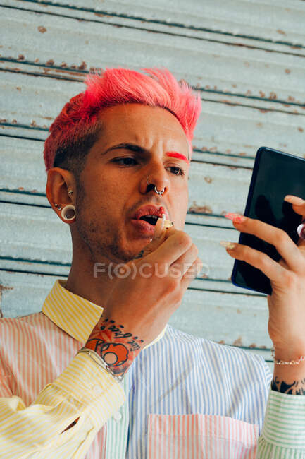 Стильный гей с татуировкой и розовыми волосами нанося помаду на губы, глядя на мобильный телефон на сером фоне — стоковое фото