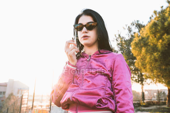 Millennial female in trendy wear and sunglasses smoking cigarette against urban building under white sky — Stock Photo