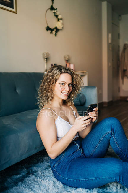 Side view of happy young female with curly blond hair in light top and jeans messaging on mobile phone and smiling while sitting on carpet in living room — Stock Photo
