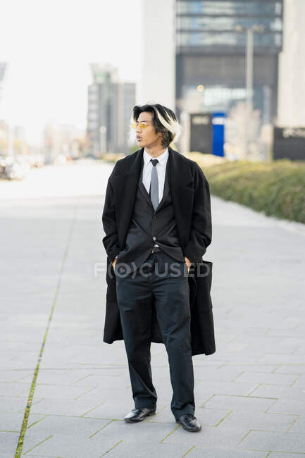 Уверенный в себе молодой этнический предприниматель мужчина в формальном костюме и пальто стоя с руками в карманах, глядя в сторону в городе — стоковое фото