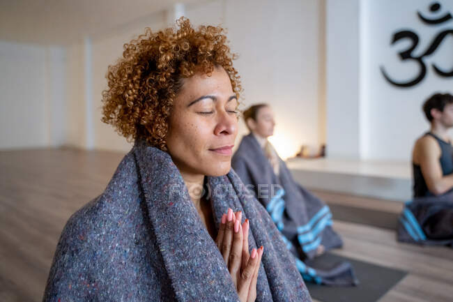 Vue latérale d'une femme ethnique enveloppée dans une couverture méditant avec des mains namastes en studio avec diverses personnes pratiquant le yoga ensemble — Photo de stock