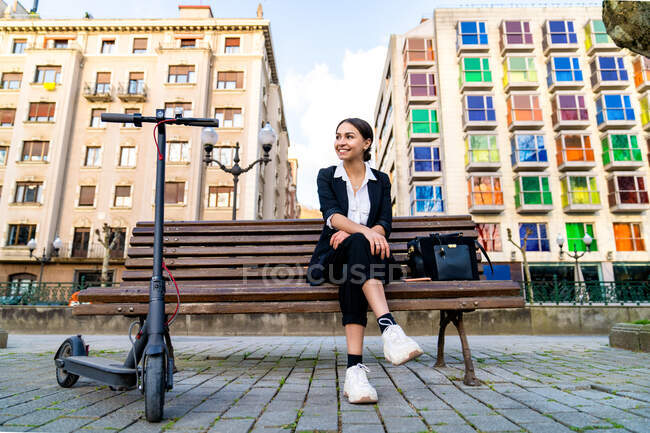 Zufriedene junge ethnische Unternehmerin sitzt mit gekreuzten Beinen auf Bank und schaut weg von Elektroroller und städtischen Gebäuden — Stockfoto