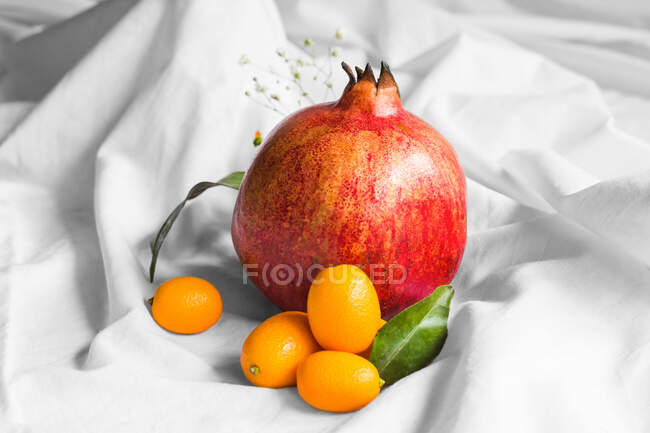 Bright whole fresh pomegranate with cumquats and leaves with sprigs on creased fabric on white background — Photo de stock