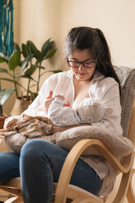 Mãe em óculos amamentando criança anônima na almofada macia enquanto sentado na sala da casa à luz do dia — Fotografia de Stock