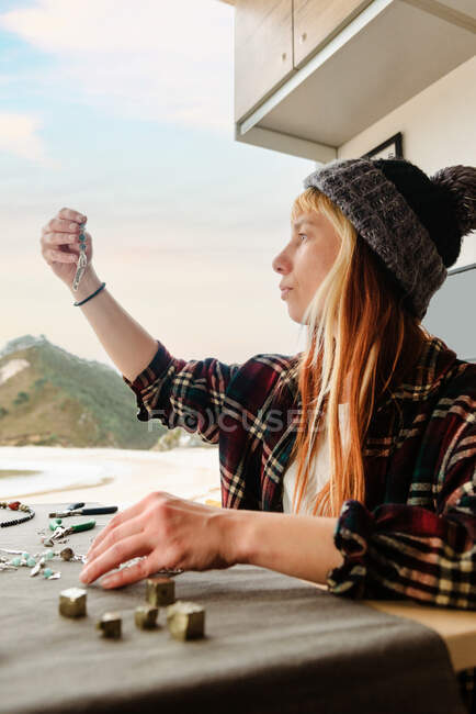 Талантливая путешествующая женщина-ремесленница примеряет браслет ручной работы, сидя за столом в грузовике на берегу моря во время путешествия — стоковое фото