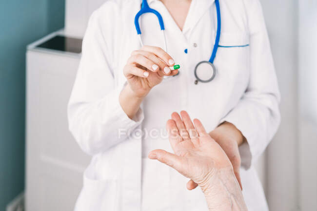 Médico femenino irreconocible en uniforme y máscara desechable que prepara medicamentos para paciente anciano anónimo durante la consulta en la clínica - foto de stock