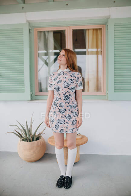 Longitud completa atractiva joven hembra en ropa de verano de pie cerca de la casa contemporánea y mirando hacia arriba - foto de stock