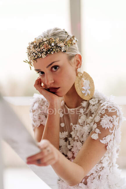 Young female in stylish bohemian white bridal dress and high heeled boots with ornamental wreath and earrings standing on stairway and looking at camera — Stock Photo