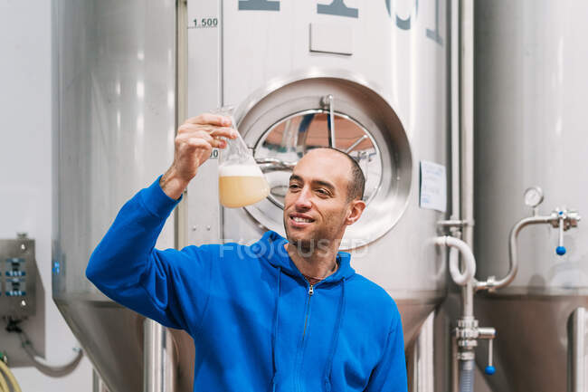 Веселий чоловічий підприємець з алкогольною рідиною в колбі працює проти бродіння цистерн в пивоварні — стокове фото