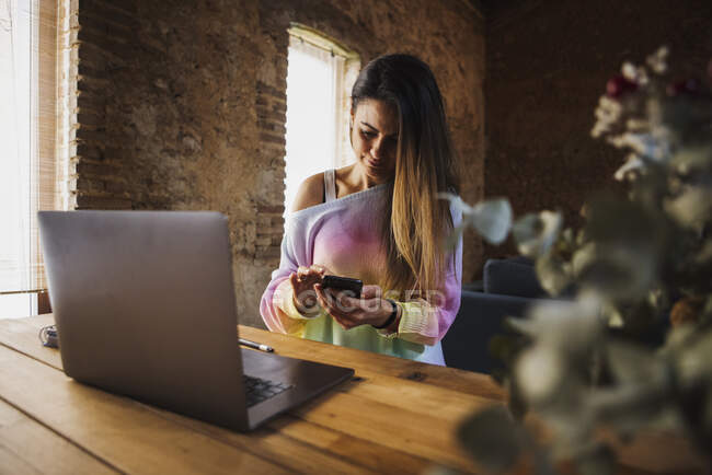 Жіноча відстань працівників текстових повідомлень на мобільному телефоні за дерев'яним столом з нетбуком у легкому будинку — стокове фото