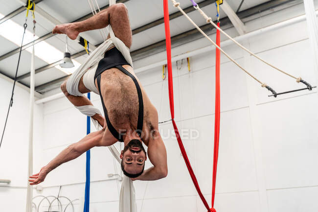 Гибкий спортсмен без рубашки, висящий вверх ногами на воздушных шелках и смотрящий в камеру во время тренировки в легком спортзале — стоковое фото