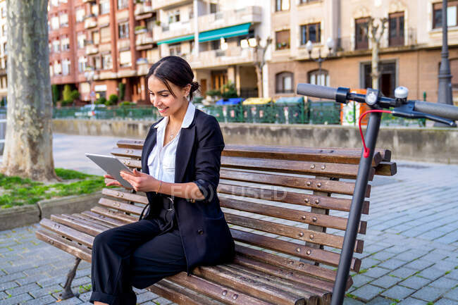 Contenido joven empresaria étnica sentada en el banco mientras navega en la tableta cerca de scooter eléctrico y edificios de la ciudad - foto de stock