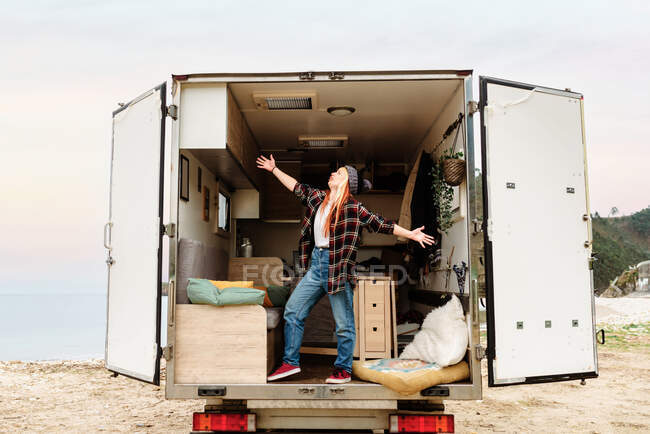 Вид сбоку мечтательной путешествующей женщины-хипстера, стоящей внутри грузовика, припаркованного у моря в горной местности и наслаждающейся отпуском с протянутыми руками — стоковое фото