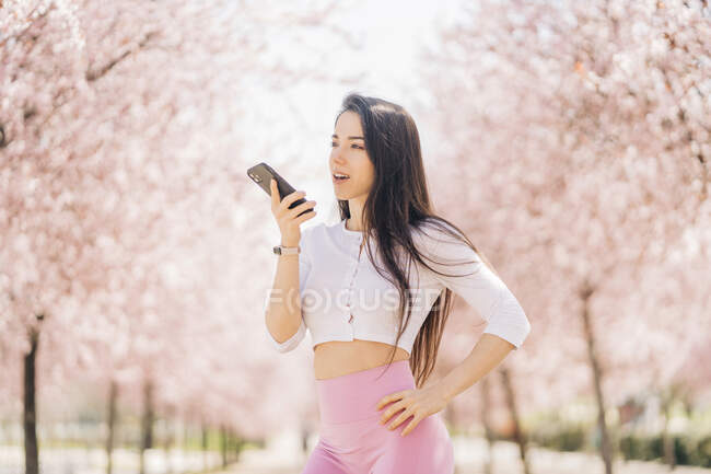 Mujer tierna joven con la mano en la cadera enviando mensaje de voz en el teléfono celular mientras mira hacia otro lado a la luz del día - foto de stock