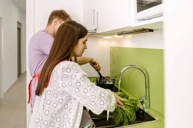 Вид сбоку на молодую этническую леди с длинными темными волосами в модной блузке, стирающую свежий зеленый салат в раковине рядом с анонимным парнем, готовящую еду в кастрюле на кухне — стоковое фото