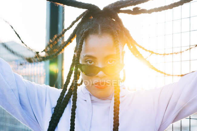 Молодая афроамериканка с красными губами и афрокосичками в солнечных очках, смотрящая на камеру под открытым небом — стоковое фото