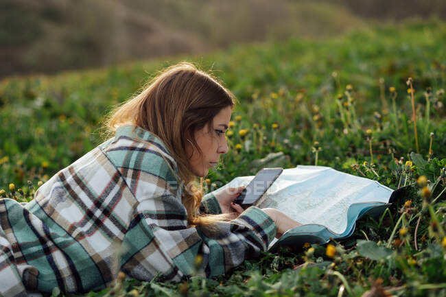 Вид збоку молодої жінки-туристки з мобільним телефоном, який дивиться карту маршруту, лежачи на траві в сільській місцевості — стокове фото