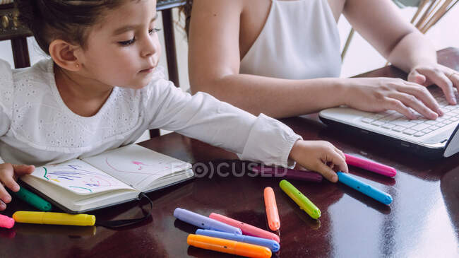 Обрізати невпізнавану жінку, яка переглядає ноутбук, поки маленька дитина сидить за столом і малює маркерами в блокноті — стокове фото