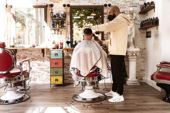 Peluquería masculina en máscara textil recortando pelo de cliente anónimo en sillón contra espejo en barbería - foto de stock