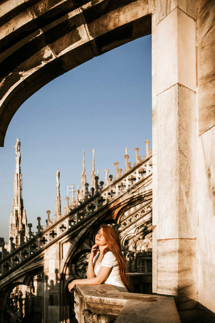 Vista lateral de una viajera irreconocible admirando Milán desde el balcón de piedra de la iglesia envejecida bajo el sol en Italia - foto de stock