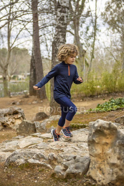 Малюк у спортивному одязі бігає на грубому камені під час тренування проти зарослих дерев у денне світло — стокове фото