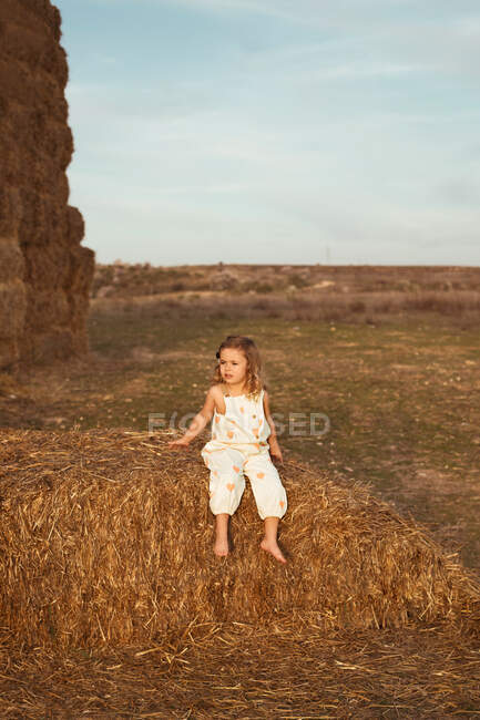 Безтурботний дитина в комбінезоні сидить на солом'яному тюку в сонячний день в сільській місцевості — стокове фото