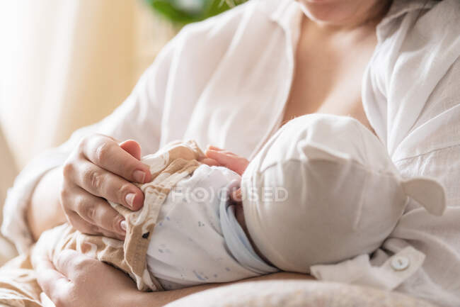 Récoltée maman méconnaissable dans des lunettes de vue allaitant petit enfant anonyme assis dans la chambre de la maison à la lumière du jour — Photo de stock