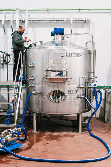 Seitenansicht eines männlichen Ingenieurs bei der Untersuchung eines Edelstahltanks mit Schläuchen in einer Brauerei mit feuchtem Boden — Stockfoto