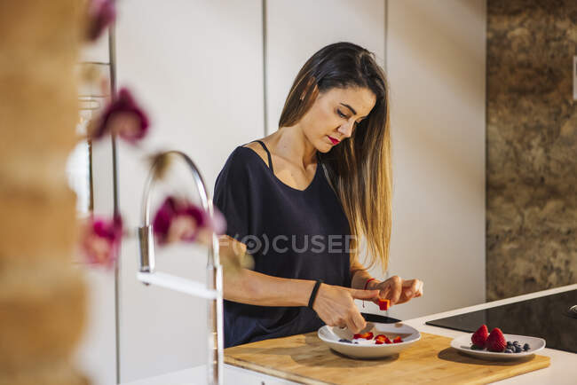 Zufriedenes Weibchen mit Messer, das reife Erdbeeren schneidet, während es zu Hause gesunde Nahrung in der Schüssel zubereitet — Stockfoto