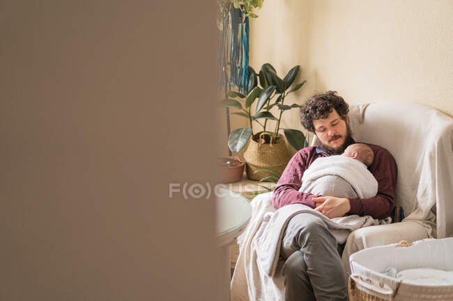 Adulto pai barbudo com pernas cruzadas e anônimo criança sentada em poltrona no quarto da casa — Fotografia de Stock