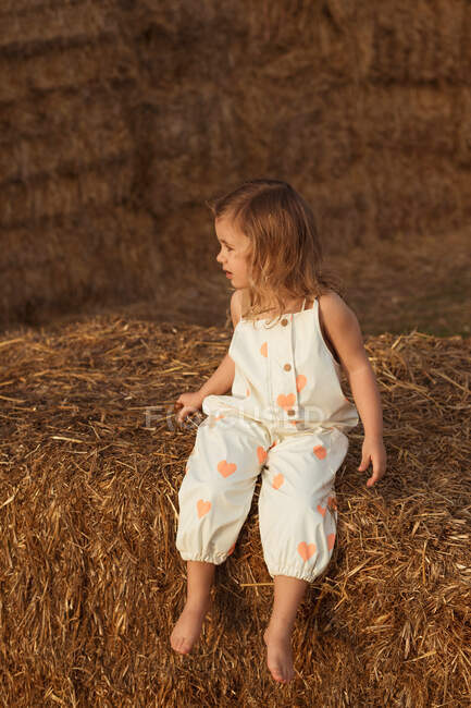 Carefree child in overalls sitting on straw bale on sunny day in countryside — Stock Photo