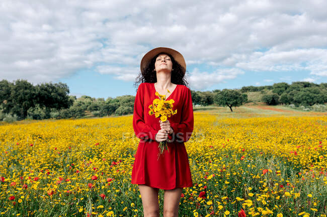 Femmina in cappello con gli occhi chiusi azienda fioritura fiori gialli nel campo di campagna sotto cielo nuvoloso — Foto stock