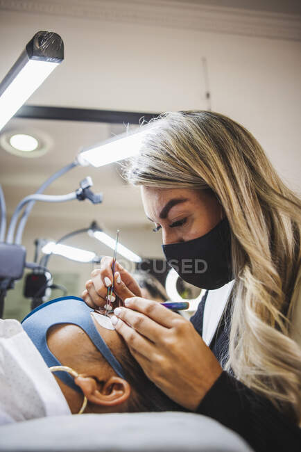 Cosmetologist with tweezers applying fake eyelashes for extension on eye of ethnic client with face protective mask in salon during coronavirus pandemic — Stock Photo