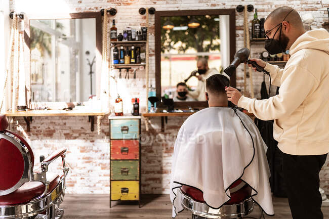 Стилист в текстильной маске с феном против мужчины в плаще в кресле в парикмахерской — стоковое фото
