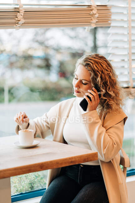 Conteúdo fêmea sentada à mesa na casa de café e mexendo bebida saborosa na xícara enquanto conversa no smartphone — Fotografia de Stock